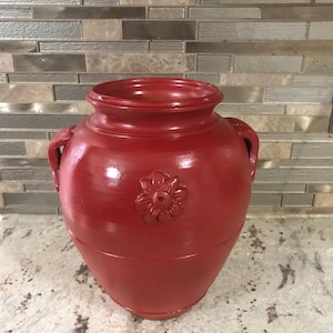 Puede incluir: Un jarrón de cerámica rojo con un diseño floral. El jarrón tiene dos asas y un cuerpo redondeado.