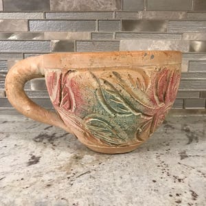 Puede incluir: Una taza de terracota con un diseño floral tallado en tonos de rosa, verde y marrón. La taza tiene un asa y está hecha de una arcilla rugosa y texturizada.