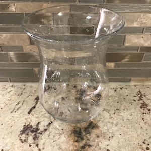 May include: A clear glass vase with a flared top and a rounded body. The vase sits on a small, clear base. The background features a neutral-toned tile backsplash and a speckled countertop.