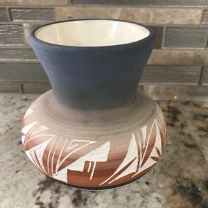 Pode incluir: Um vaso de cerâmica artesanal com uma borda larga azul escuro e um corpo bulboso. O vaso apresenta um gradiente de cores, passando do castanho claro ao castanho avermelhado, com padrões geométricos brancos. O interior é branco.