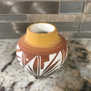 Peut inclure: Petit vase rond de couleur terre cuite avec un intérieur blanc et un bord jaune. Le vase présente un motif géométrique blanc autour du corps. Le vase est probablement fait à la main et adapté à des fins décoratives.