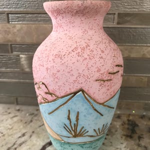 Peut inclure: Un vase décoratif avec une partie supérieure rose mouchetée et une scène de montagne bleue et dorée sur la moitié inférieure. Le vase a une base vert clair et des accents dorés. Le vase est probablement en céramique.