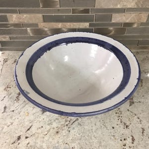 May include: A white ceramic bowl with a blue rim and a blue stripe near the edge. The bowl is round and has a speckled interior. The bowl is sitting on a speckled countertop.