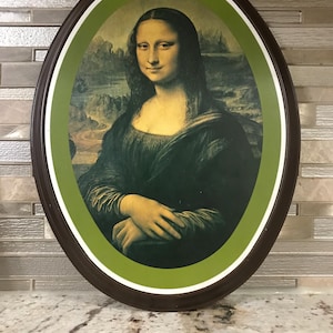 Könnte beinhalten: Ovales Dekotablett mit dem Gemälde der Mona Lisa. Das Kunstwerk ist mit einem braunen Rand, einem weißen Innenrand und einem grünen Hintergrund gerahmt. Ein Vintage-Artikel.