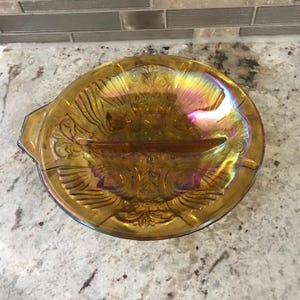 May include: An iridescent amber-coloured glass serving dish with a divided section. The dish has an ornate, embossed floral and scroll design. The dish has a handle on each side and measures approximately 25 cm in diameter.