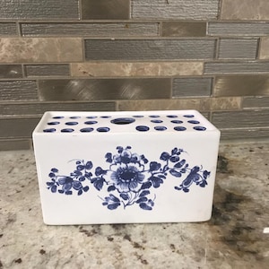 May include: A white rectangular ceramic flower frog with a blue floral design. The frog has multiple holes on top for arranging flowers. The design features a large flower and leaves in a classic blue and white color scheme.