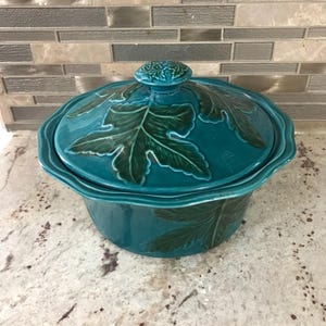 May include: Teal ceramic casserole dish with a lid. The dish is decorated with green leaf designs.