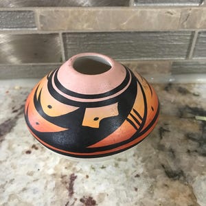 Może przedstawiać: Mały, okrągły ceramiczny wazon z wąskim otworem. Wazon ma wzór czarnych, pomarańczowych i brzoskwiniowych kształtów geometrycznych. Podstawa jest biała. Wazon jest prawdopodobnie ręcznie robiony i nadaje się do dekoracji wnętrz.