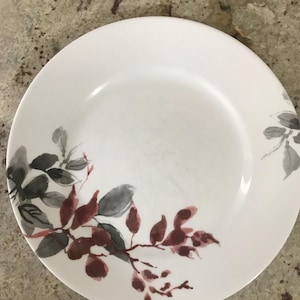 May include: White ceramic plate with a watercolor design of leaves and branches in shades of gray and burgundy. The plate has a simple, elegant design, suitable for serving food.