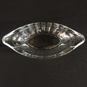 Puede incluir: Un plato de servir de vidrio transparente con forma ovalada redondeada y dos asas. El plato tiene un diseño texturizado y acanalado.