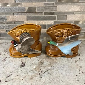 以下が含まれることがあります： 茶色と白の鳥と青と緑の鴨がついた、セラミック製のブーツ型のプランター2つ。ブーツはどちらも茶色で、バックルが付いています。