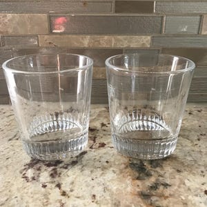 May include: Two clear, short drinking glasses with a textured base. The glasses are cylindrical with a slightly flared rim. They are displayed on a speckled countertop, with a neutral-toned tile backsplash in the background. Ideal for serving beverages.