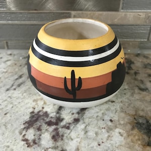 Può includere: Un vaso rotondo in ceramica con un design a paesaggio desertico. Il vaso presenta una parte superiore gialla con strisce nere e bianche e una scena dipinta di un cactus e un tramonto nelle tonalità del marrone e dell'arancione.