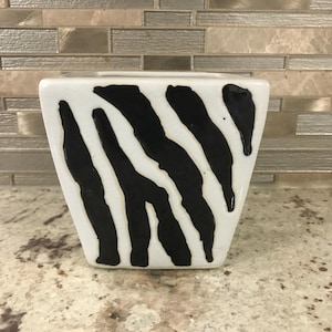 以下が含まれることがあります： 白地に黒のゼブラ柄が施されたセラミック製の花瓶。花瓶は四角い形で、底に向かってわずかに細くなっています。黒い縞模様は太く不規則で、白い背景に対して大胆なコントラストを生み出しています。
