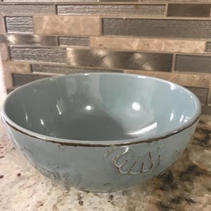 May include: A pale blue ceramic bowl with a glossy finish. The bowl features a brown rim and embossed seashell designs. This decorative bowl is suitable for serving food or as a kitchen accent.