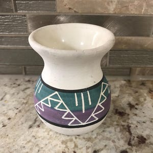 Könnte beinhalten: Kleine, weiße Keramikvase mit ausgestelltem Rand und rundem Körper. Die Vase hat ein geometrisches Design in Türkis, Lila und Weiß mit schwarzen Akzenten. Die Vase ist wahrscheinlich handgefertigt und hat eine rustikale, handwerkliche Ausstrahlung.