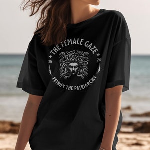 Puede incluir: Camiseta negra con un gr&aacute;fico blanco de la cabeza de Medusa rodeada de serpientes. El gr&aacute;fico est&aacute; en un c&iacute;rculo con el texto "The Female Gaze" y "Petrify the Patriarchy" alrededor.