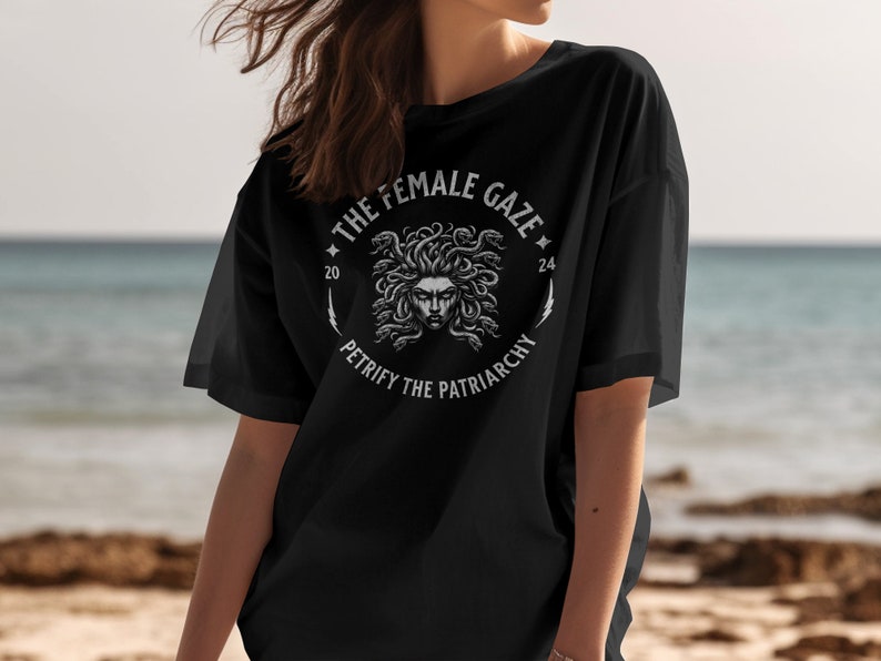 Puede incluir: Camiseta negra con un gr&aacute;fico blanco de la cabeza de Medusa rodeada de serpientes. El gr&aacute;fico est&aacute; en un c&iacute;rculo con el texto "The Female Gaze" y "Petrify the Patriarchy" alrededor.