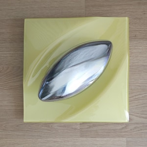 Könnte beinhalten: Eine quadratische, gelbe Glasplatte mit einem silbernen, bootsförmigen Objekt in der Mitte. Die Platte hat eine glänzende Oberfläche. Das silberne Objekt reflektiert das Licht. Auf einer Holzoberfläche.