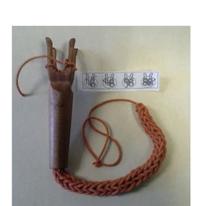 Puede incluir: Una herramienta de tejido de madera con un mango largo y tres puntas en la parte superior. Un hilo naranja está unido a la herramienta y se está utilizando para crear un cordón tejido. Un diagrama del proceso de tejido se muestra en un trozo de papel.