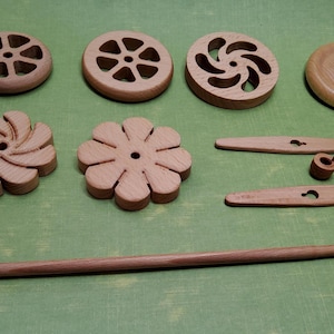 Puede incluir: Un conjunto de ruecas de madera para hilo, con diferentes diseños, incluyendo una flor, una espiral y una estrella. El conjunto también incluye dos clavijas de madera y un huso de madera.