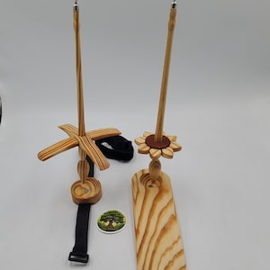 May include: Two wooden spinning wheels with a black strap and a wooden base. The spinning wheel on the left has a four-pronged design and the spinning wheel on the right has a flower-shaped design.