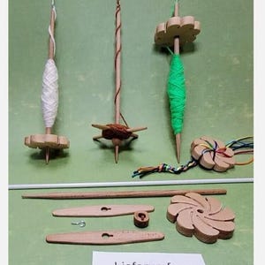 Könnte beinhalten: Drei Holzspinnräder mit unterschiedlich farbigem Garn, sowie eine Holzspindel und zwei Holzstift. Der Text "Lieferumfang" ist im Bild sichtbar.