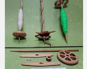 May include: Three wooden spinning wheels with different colored yarn, a wooden dowel, two wooden spindles, and a wooden wheel. The text "Lieferumfang" is written on a white card.