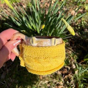 Könnte beinhalten: Eine Keramiktasse mit einem froschartigen Design, gelben Augen und einem gestrickten gelben Überzug. Die Tasse wird in einer Hand gehalten, mit Narzissen im Hintergrund. Die Tasse hat ein einzigartiges, handgefertigtes Aussehen.