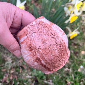 Può includere: Piccolo vaso in ceramica rosa e marrone fatto a mano, con superficie strutturata. Il vaso è tenuto in mano, con fiori ed erba sfocati sullo sfondo. Il colore del vaso è un mix di rosa e marrone.