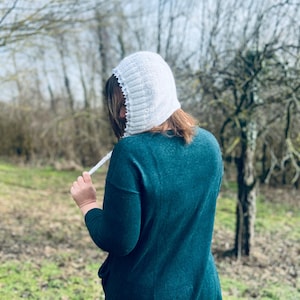 Könnte beinhalten: Eine weiße Strickhaube mit gewelltem Rand und Bindebändern. Die Haube wird über einem teal-grünen Langarm-Top getragen. Der Hintergrund zeigt kahle Bäume und grünes Gras.