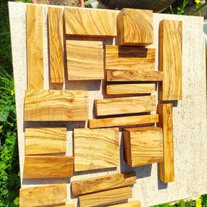 May include: A collection of various-sized olive wood blocks, showcasing natural grain patterns and warm tones. Some blocks have dimensions written on them in inches. Ideal for woodworking projects or crafts.
