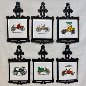 May include: Set of six vintage-style trivets with black cast iron frames and ceramic tiles. Each tile features a colorful illustration of a classic car with the car's name and year. The trivets have a decorative top with a turquoise accent.