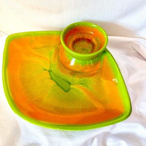 May include: A ceramic serving dish featuring a large, asymmetrical, leaf-shaped tray with a smaller, circular bowl attached to one side. The piece has a vibrant, glossy glaze in blended shades of bright lime green and warm orange throughout.