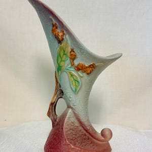 May include: A ceramic cornucopia vase featuring a matte finish with a gradient of dusty rose at the base transitioning to a pale blue upper section. The piece is decorated with raised, molded brown branches and green leaves on the side.