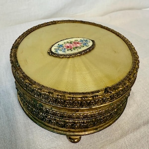 May include: A round, gold-tone trinket box with a raised, oval-shaped design on the lid. The design features a floral motif with pink, blue, and white flowers.