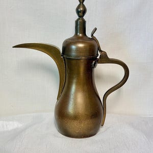 May include: A vintage brass coffee pot with a long spout and curved handle. The pot has a bulbous body and a decorative finial on the lid. The brass has a warm, aged patina, suggesting it is an antique or vintage item.