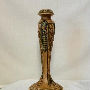 May include: A tall, tan ceramic candlestick holder featuring a flared base and a slender, tapered stem. The surface is decorated with vertical, textured leaf-like carvings and a small, colorful floral motif near the top rim, finished in a glossy glaze.