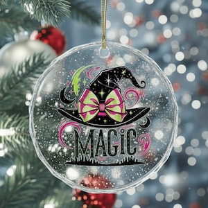 Könnte beinhalten: Runder, klarer Weihnachtsschmuck mit einem schwarzen Hexenhut-Design, einer rosa und grünen Schleife und dem Wort "MAGIC". Der Schmuck hängt an einem goldenen Faden.