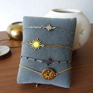 Könnte beinhalten: Vier zarte Armbänder mit himmlischen Anhängern werden auf einem grauen Kissen präsentiert. Die Armbänder zeigen Stern-, Sonnenstrahlen- und Kompass-Designs in Silber- und Goldtönen. Das Kompassarmband hat einen runden Anhänger mit einer Windrose.
