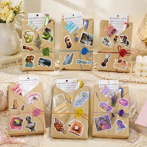 Puede incluir: Bolsas de regalo de papel kraft decoradas con varias pegatinas, incluyendo un autobús vintage, una mariposa y querubines. Cada bolsa contiene una tarjeta de "READING LIST" y está atada con hilo y un adorno decorativo. Se ven bolígrafos y artículos de papelería.