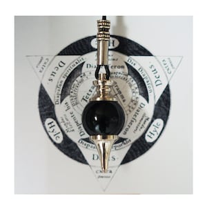 May include: A silver pendulum with a black, round bob hangs from a black cord. The pendulum is suspended over a circular chart with the words "Deus", "Hyle", "Diapafon", "Tetra", "Diapente", "Diateſseron", and "Diapas" printed on it.
