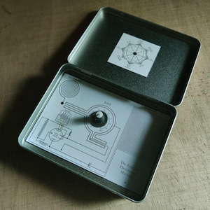 May include: A silver metal box with a hinged lid. The box contains a white paper with a diagram of a circuit and the text "The Symbolic Harmonic Machine".