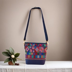 Puede incluir: Un bolso bandolera de lona azul con un estampado geométrico colorido. El bolso tiene una correa larga ajustable y un cierre de cremallera.