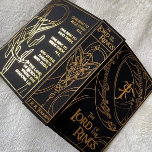 May include: A set of three black hardcover books with gold foil lettering and illustrations. The books are titled "The Lord of the Rings" and feature quotes from the book, including "One Ring to rule them all, One Ring to find them, One Ring to bring them all, and in the darkness bind them."