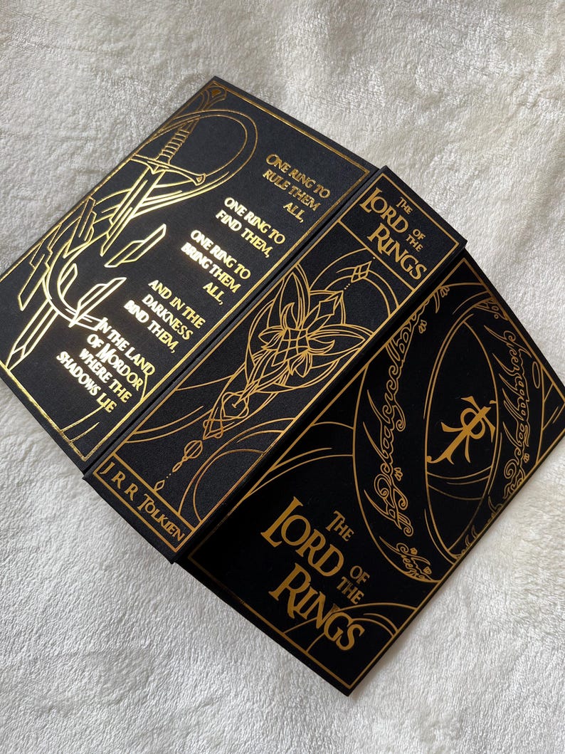 May include: A set of three black hardcover books with gold foil lettering and illustrations. The books are titled "The Lord of the Rings" and feature quotes from the book, including "One Ring to rule them all, One Ring to find them, One Ring to bring them all, and in the darkness bind them."
