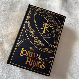 May include: A black hardcover book with a gold embossed design of the One Ring from "The Lord of the Rings" on the front cover. The title "The Lord of the Rings" is also embossed in gold.