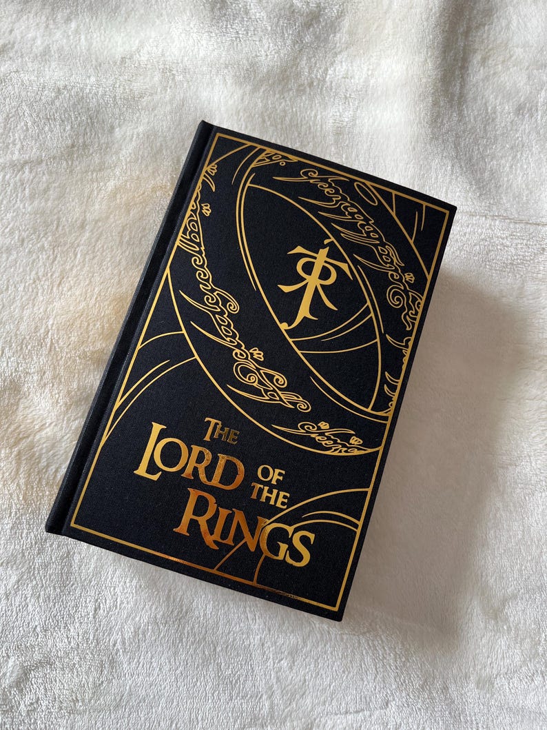 May include: A black hardcover book with a gold embossed design of the One Ring from "The Lord of the Rings" on the front cover. The title "The Lord of the Rings" is also embossed in gold.