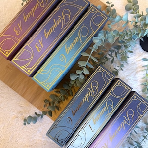 May include: Six decorative boxes with book-like designs. Each box features a different color scheme, including purple, blue, and teal, with gold lettering and accents. The boxes are labeled with words like "Binding" and "Keeping".