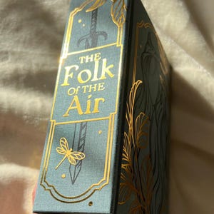 Puede incluir: Un libro de tapa dura azul verdoso con letras e ilustraciones doradas. El t&iacute;tulo es "The Folk of the Air".